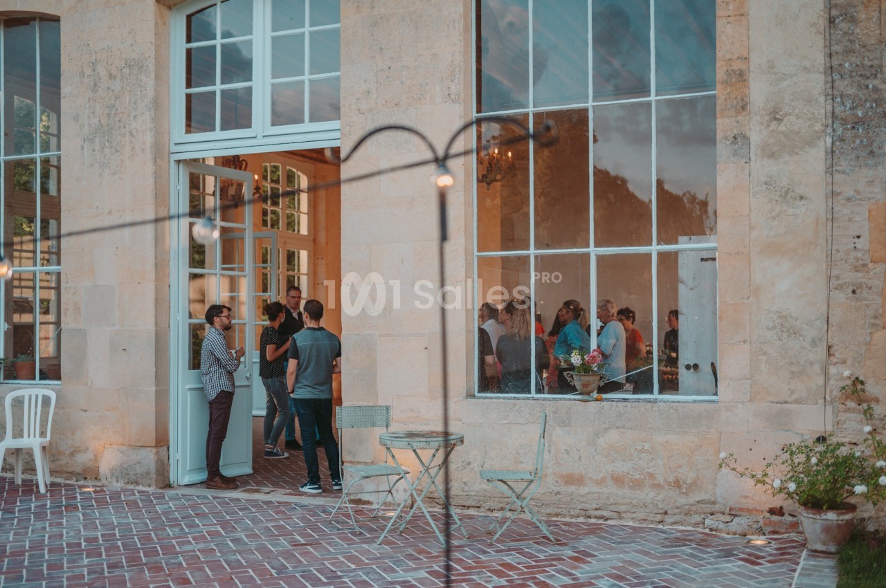 Groupe de personnes discutant à l'entrée d'un bâtiment en pierre avec de grandes fenêtres, tables et chaises en extérieur.