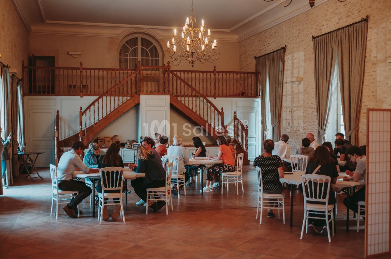 Personnes assises en petits groupes autour de tables dans une grande salle avec un escalier en bois et un lustre.