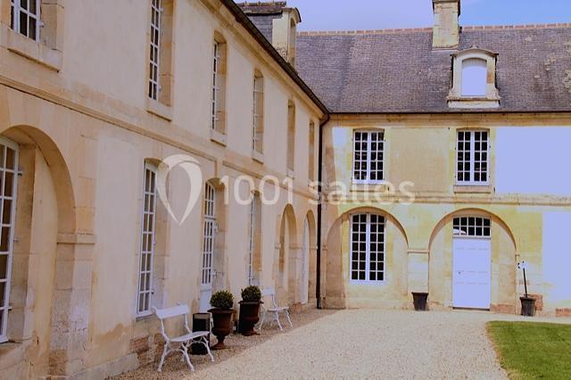 Cour intérieure avec façade en pierre claire, fenêtres à petits carreaux, bancs blancs et pots de fleurs.