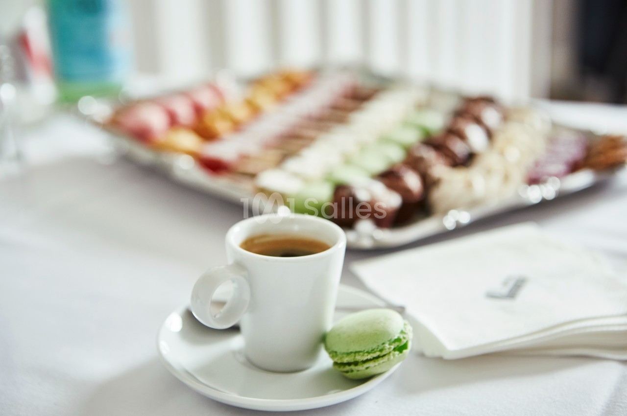 Tasse de café avec un macaron vert au premier plan, plateau de pâtisseries flou en arrière-plan.