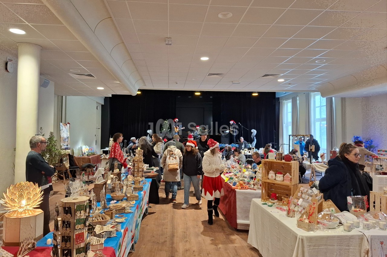 Salle animée avec des stands d'artisanat, des visiteurs et une scène décorée pour un marché de Noël.