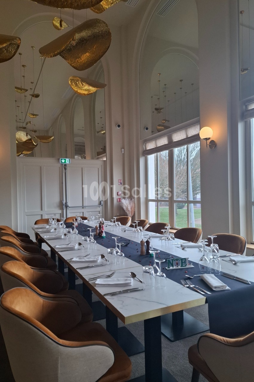 Salle de restaurant élégante avec une grande table dressée, chaises en velours et décorations dorées suspendues.