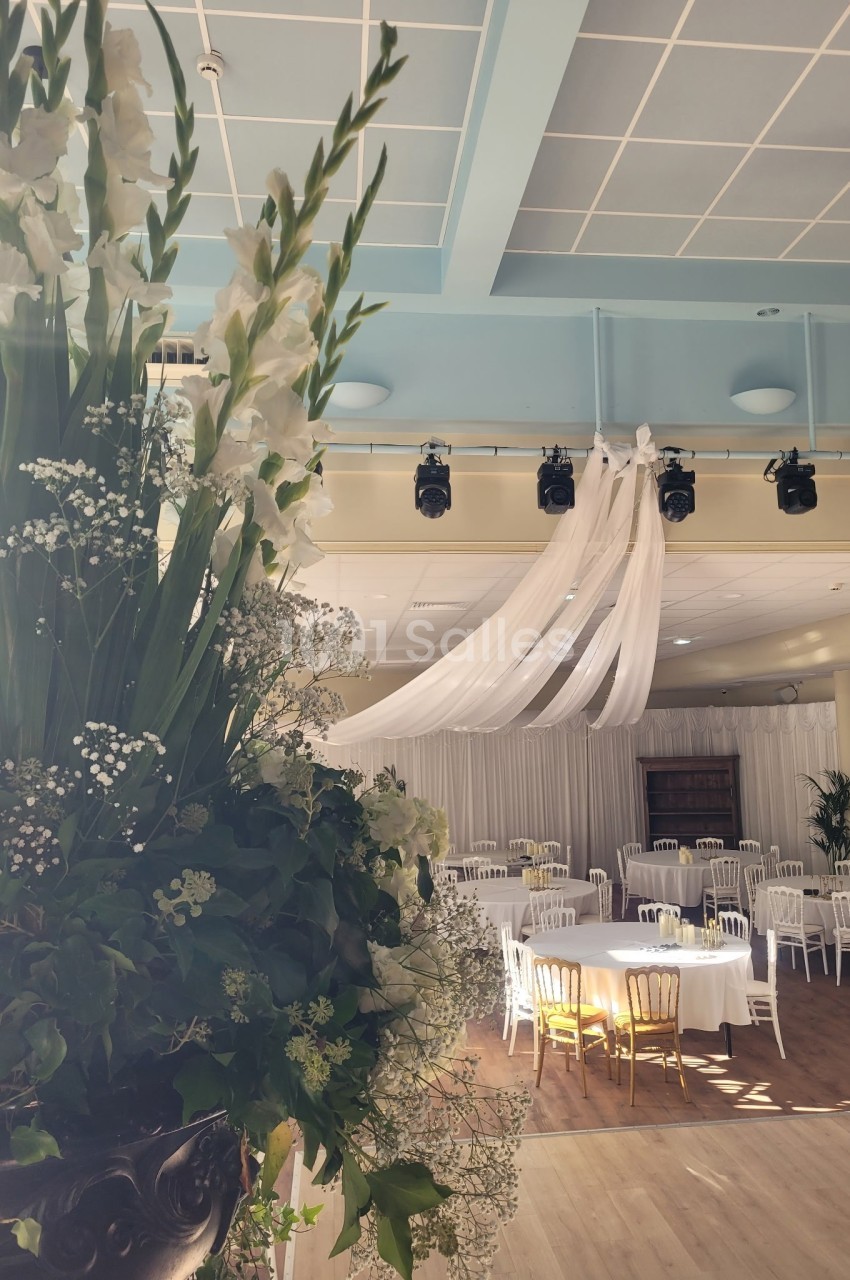 Salle de réception lumineuse avec tables rondes dressées, chaises blanches et grande composition florale au premier plan.