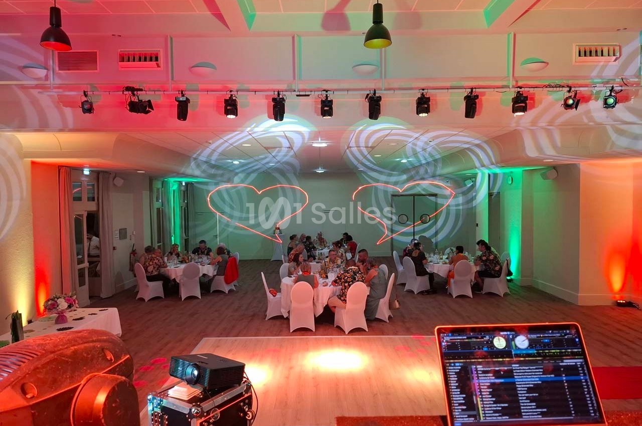 Salle de réception décorée avec des lumières colorées, des invités assis à des tables rondes et une console de DJ au premier…