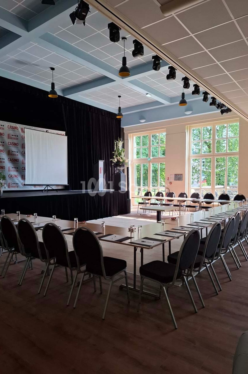 Salle de conférence lumineuse avec tables disposées en U, écran de projection et grandes fenêtres donnant sur l'extérieur.