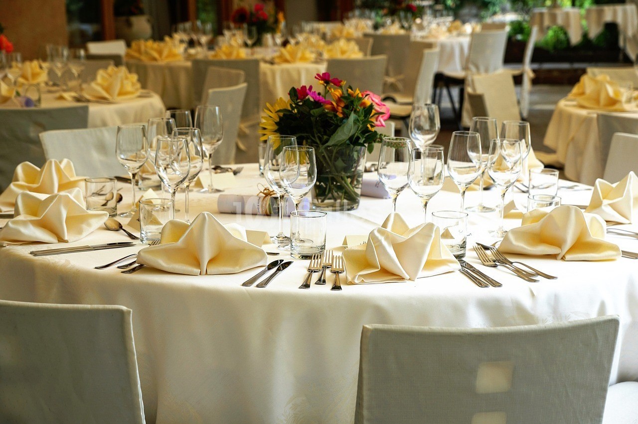 Tables élégamment dressées avec nappes blanches, serviettes pliées, verres et couverts, décorées de fleurs colorées.