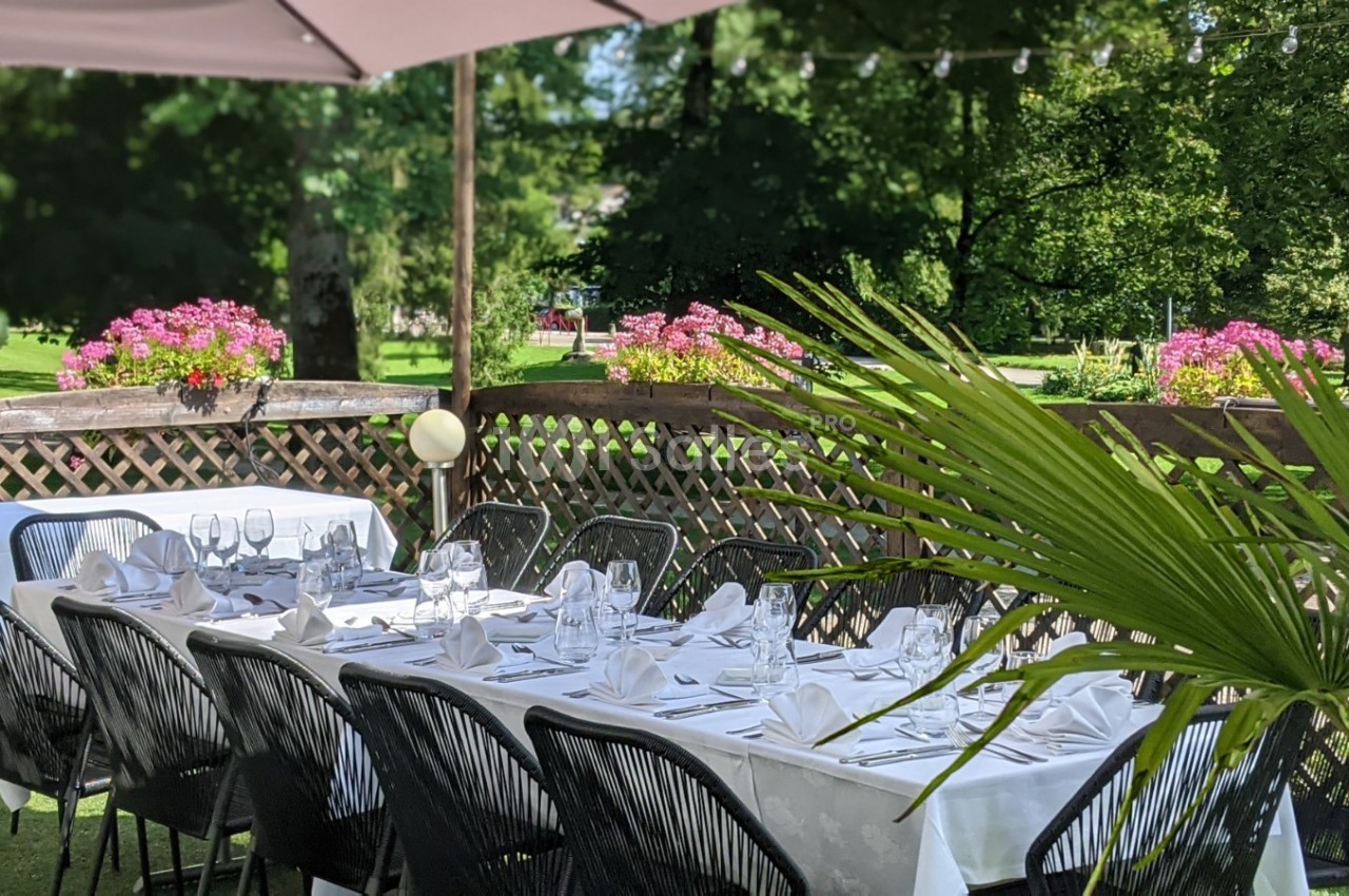 Tables dressées avec nappes blanches et chaises noires sur une terrasse ombragée, entourée de verdure et de fleurs.