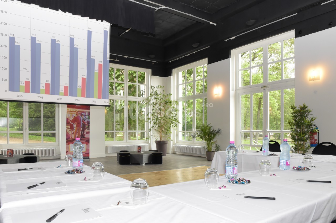 Salle de réunion lumineuse avec grandes fenêtres, tables dressées, graphiques projetés et plantes en arrière-plan.