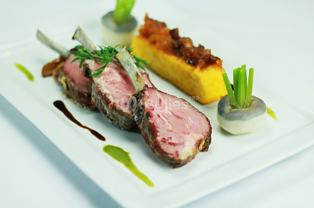 Carré d'agneau rôti avec garniture de légumes, polenta et sauces décoratives sur une assiette blanche.