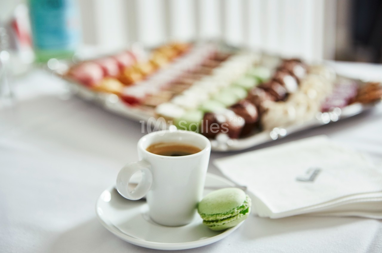 Tasse de café sur une table avec un macaron vert et un plateau de desserts flous en arrière-plan.