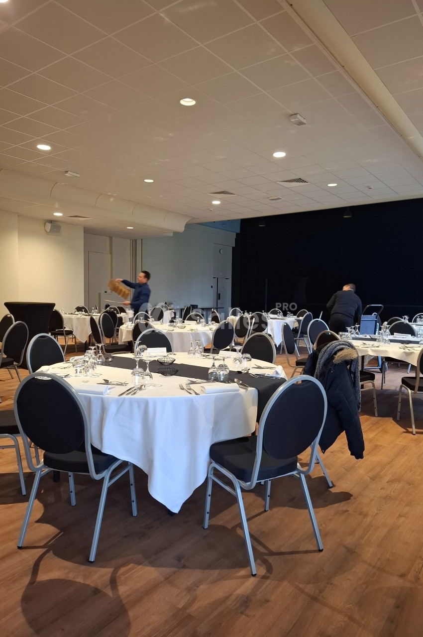 Salle de réception avec tables rondes dressées, nappes blanches et chaises, deux personnes en préparation.
