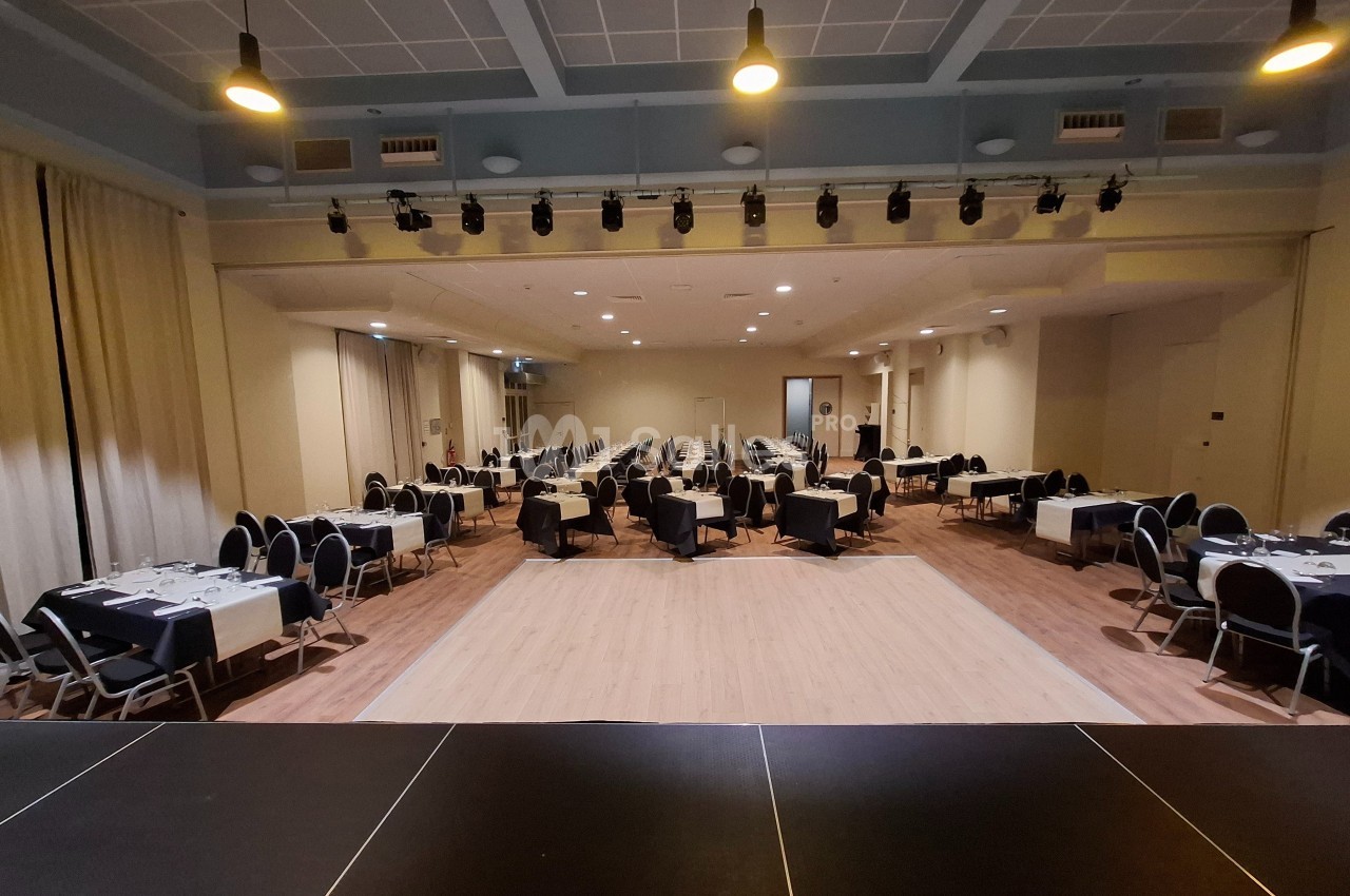 Salle de réception avec tables dressées et chaises, vue depuis une scène, éclairage suspendu au plafond.