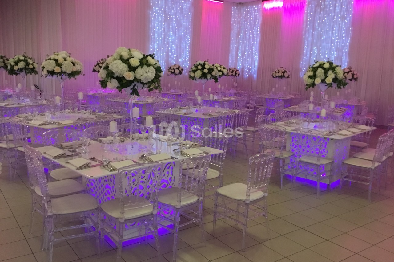 Salle de réception décorée avec des tables ornées de fleurs blanches et des chaises transparentes sous un éclairage violet.