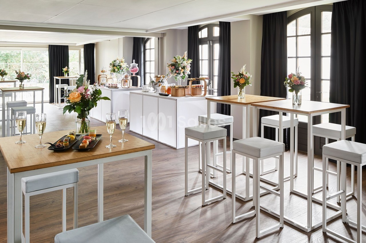 Salle lumineuse aménagée pour un événement, avec tables hautes, chaises, fleurs et buffet de boissons et amuse-bouches.
