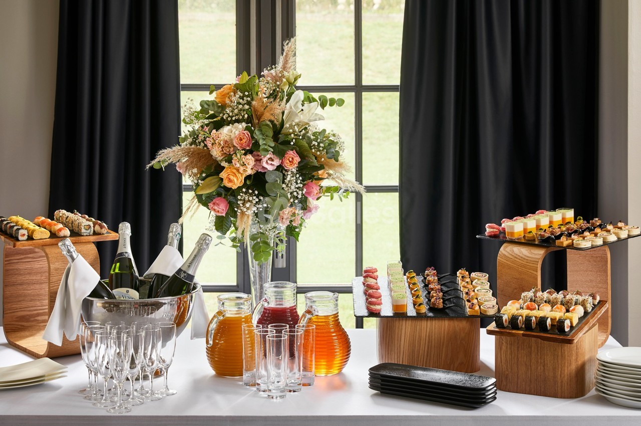 Table dressée avec des amuse-bouches variés, un bouquet de fleurs, des boissons et des verres devant une fenêtre.