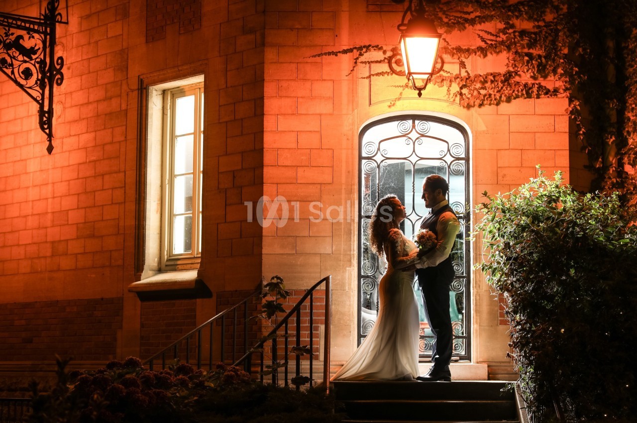 Un couple en tenue de mariage se tient devant une porte illuminée, entouré de briques et de végétation.