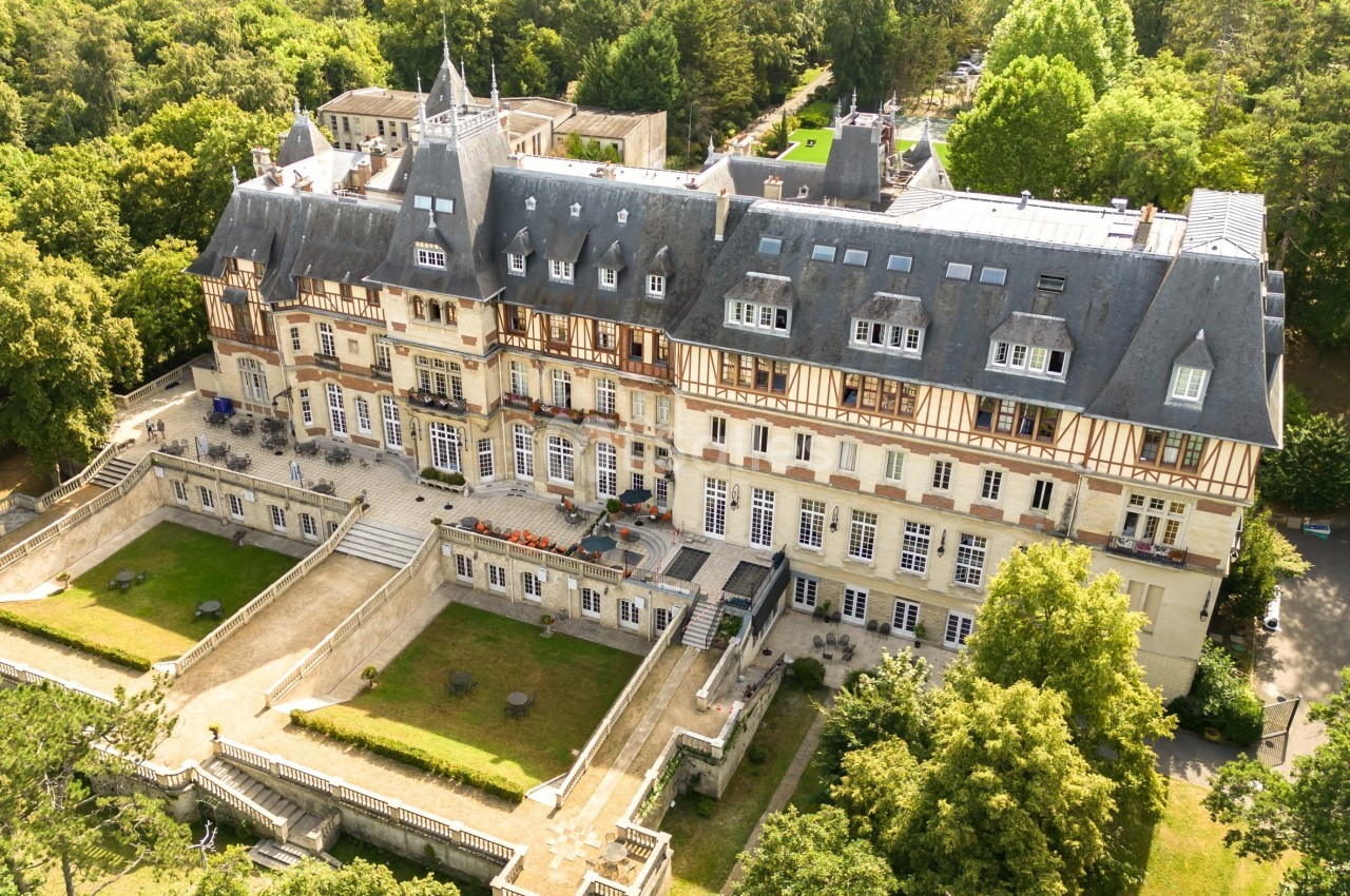 Vue aérienne d'un grand bâtiment historique entouré de jardins et d'arbres, avec une architecture à colombages.