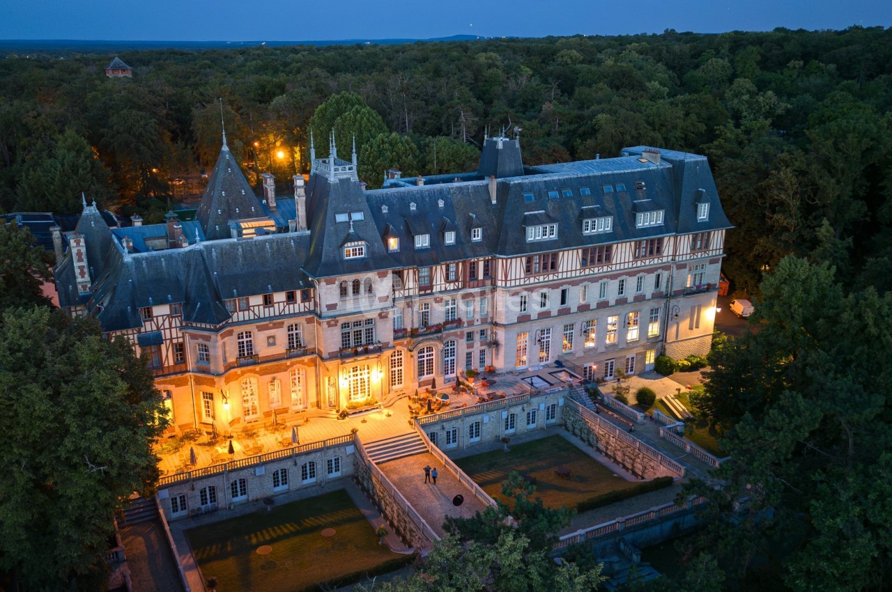 Vue aérienne d'un grand château éclairé au crépuscule, entouré de forêt dense.