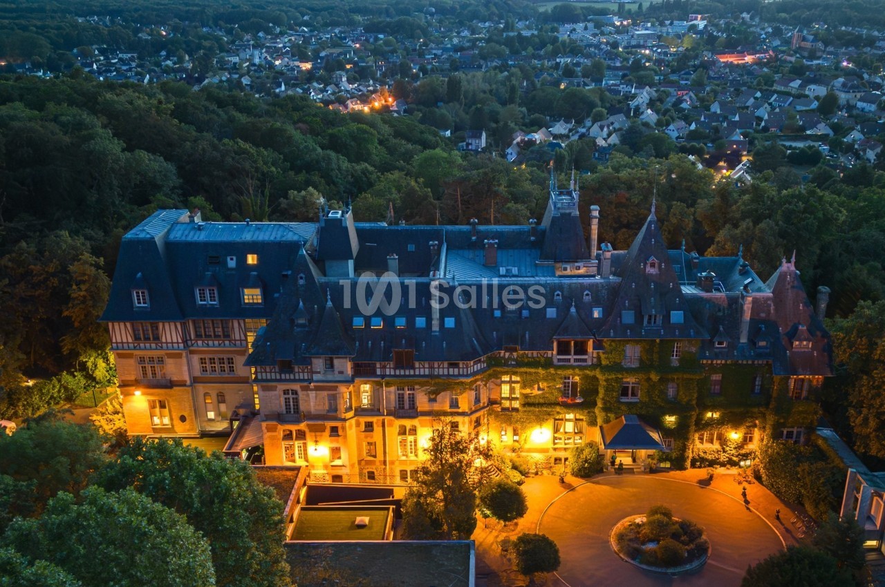Vue aérienne d'un grand bâtiment historique éclairé, entouré de forêt et surplombant une ville en soirée.