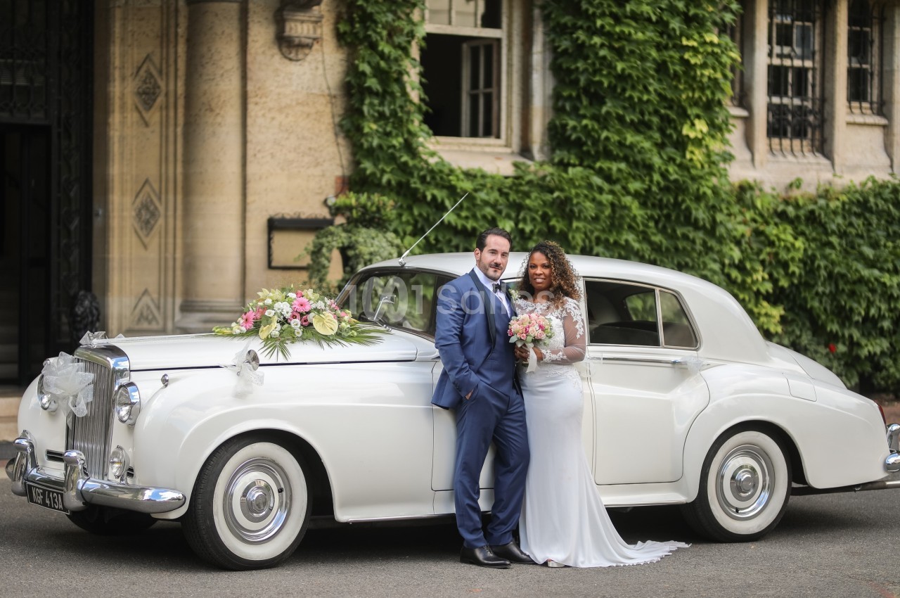Un couple en tenue de mariage pose devant une voiture ancienne blanche décorée de fleurs, près d'un bâtiment couvert de…
