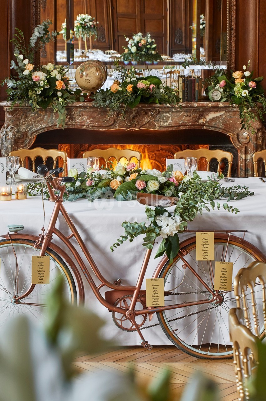 Vélo décoratif en métal cuivré orné de fleurs et de feuillage, placé devant une table de réception élégante.