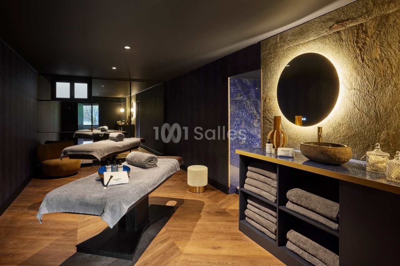 Salle de spa avec deux tables de massage, serviettes pliées, miroir rond éclairé et décor en pierre naturelle.