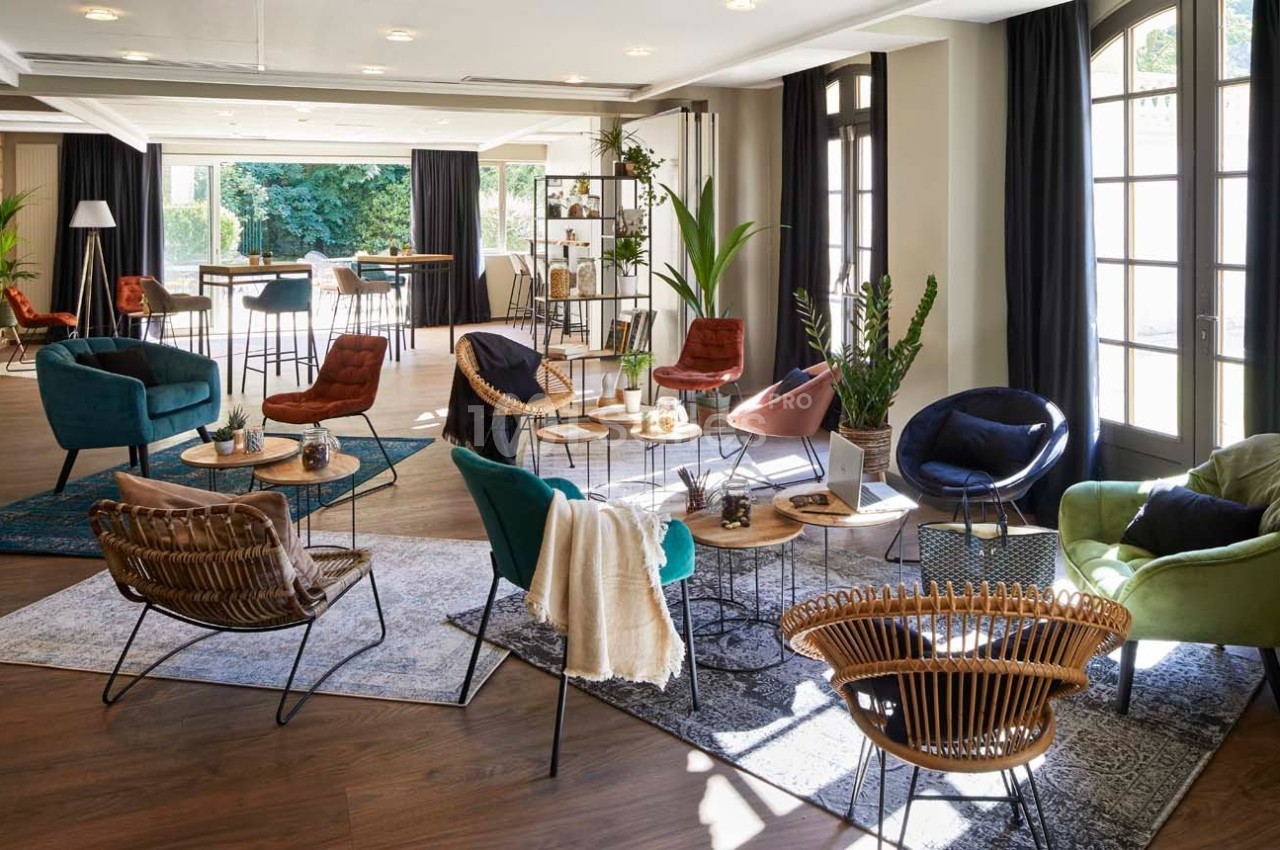 Salle lumineuse aménagée avec des fauteuils variés, des tables basses et des plantes, donnant sur un espace extérieur…