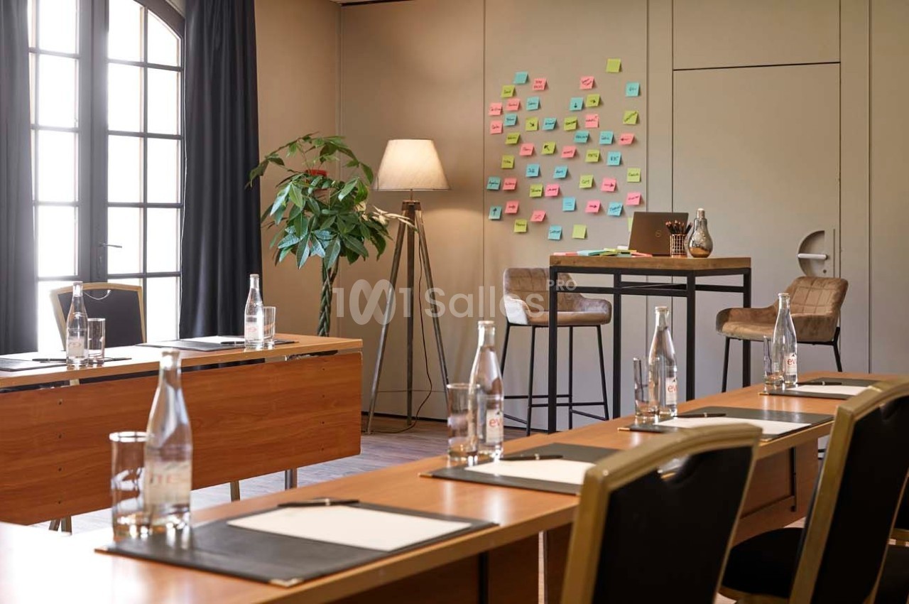 Salle de réunion lumineuse avec tables en bois, chaises noires, tableau couvert de post-it colorés et bouteilles d'eau.