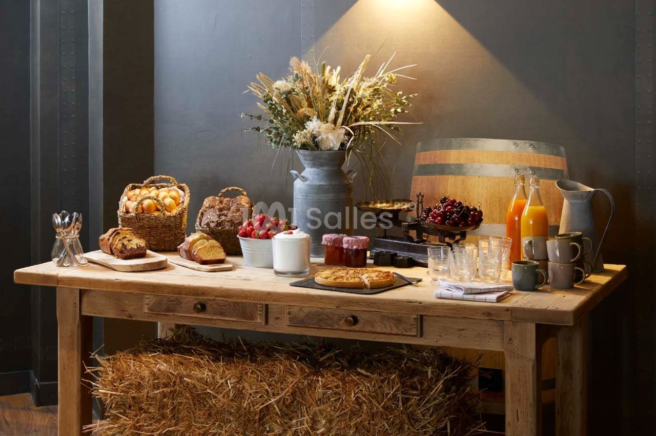 Table en bois rustique avec pains, viennoiseries, fruits, jus, confitures et fleurs séchées sous un éclairage chaleureux.