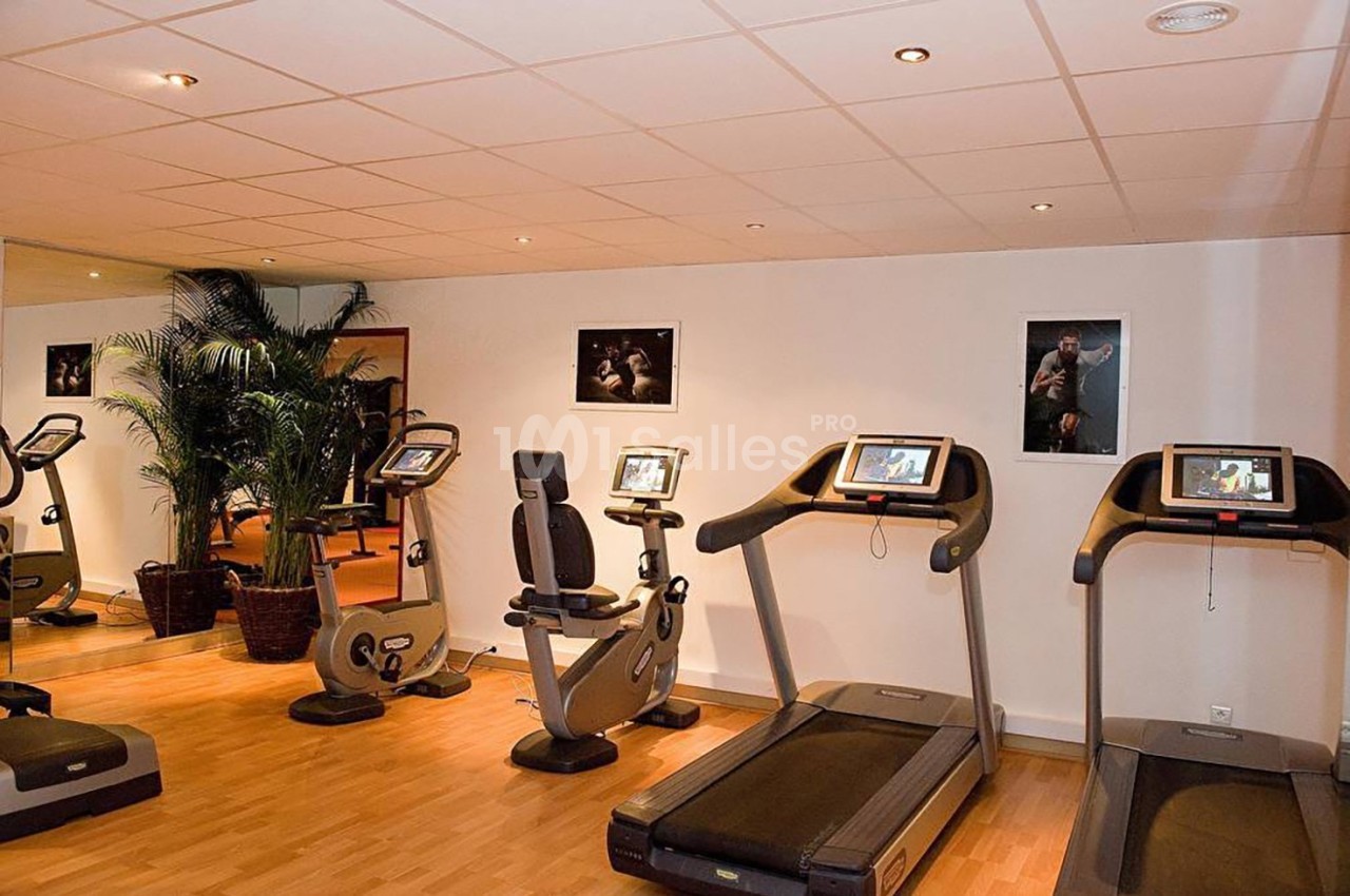 Salle de sport avec tapis de course, vélos d'exercice, plantes décoratives et affiches sur les murs.