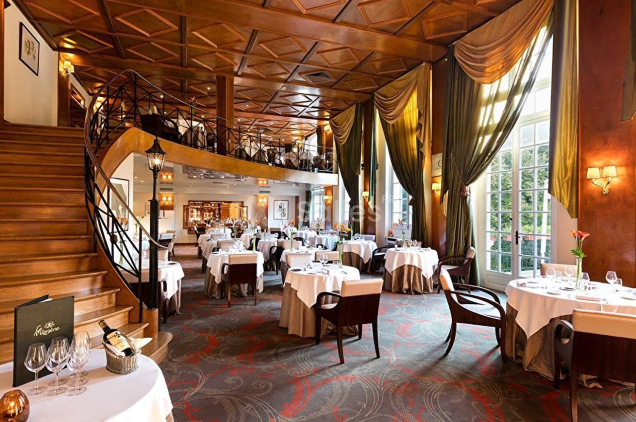 Salle de restaurant élégante avec tables dressées, escalier en bois, grandes fenêtres et rideaux longs.