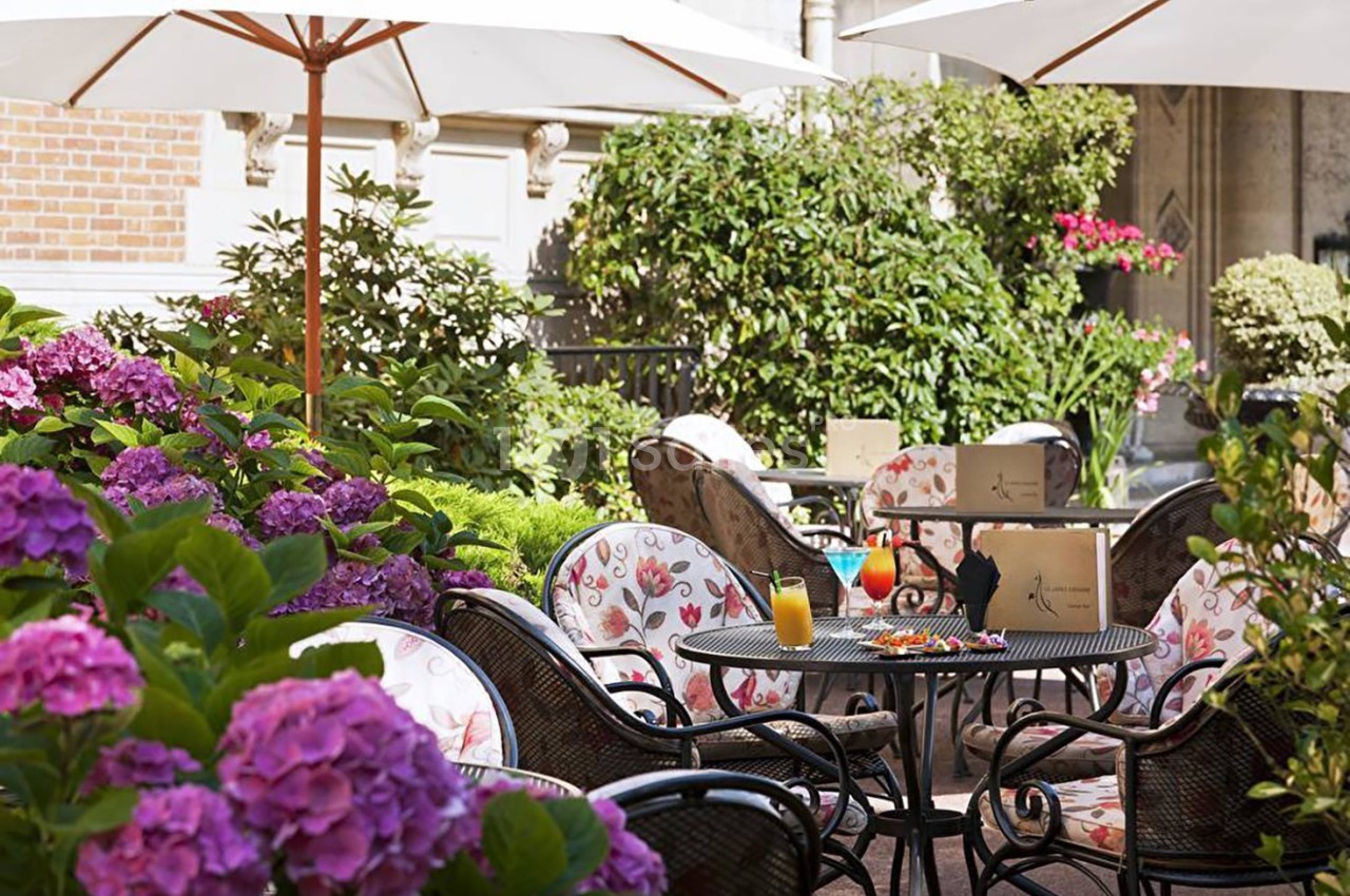 Terrasse fleurie avec tables et chaises en fer forgé, boissons servies sur une table, parasols et verdure environnante.