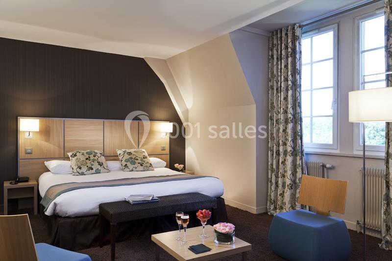 Chambre d'hôtel avec lit double, table basse, fauteuil, éclairage doux et grande fenêtre avec rideaux fleuris.