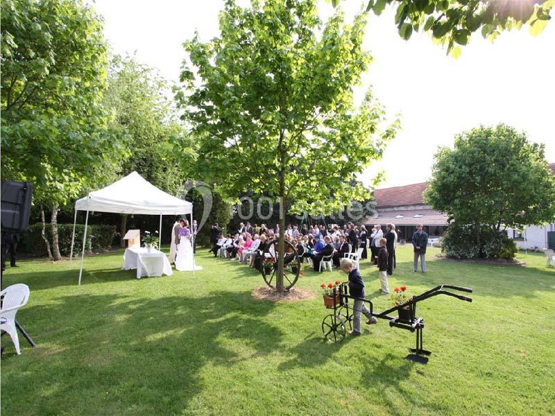Cérémonie en plein air avec des invités assis sous des arbres, une tente blanche et un espace vert environnant.