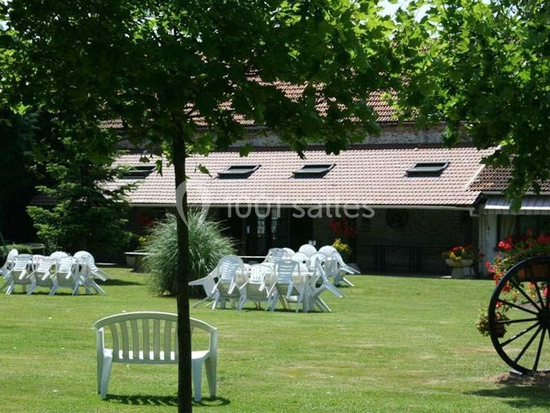 Chaises et tables blanches disposées sur une pelouse devant un bâtiment en pierre avec toit en tuiles, entouré de verdure.