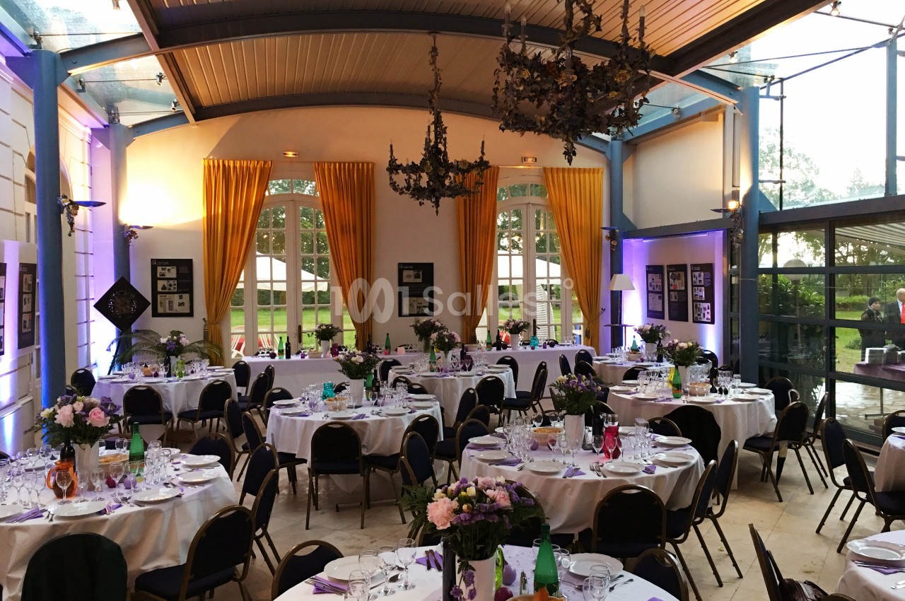Salle de réception élégante avec tables rondes dressées, décor floral et grandes fenêtres laissant entrer la lumière…
