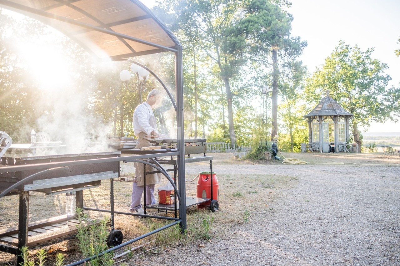 Un homme cuisine sur un barbecue en plein air sous un abri, avec un jardin et un kiosque en arrière-plan.