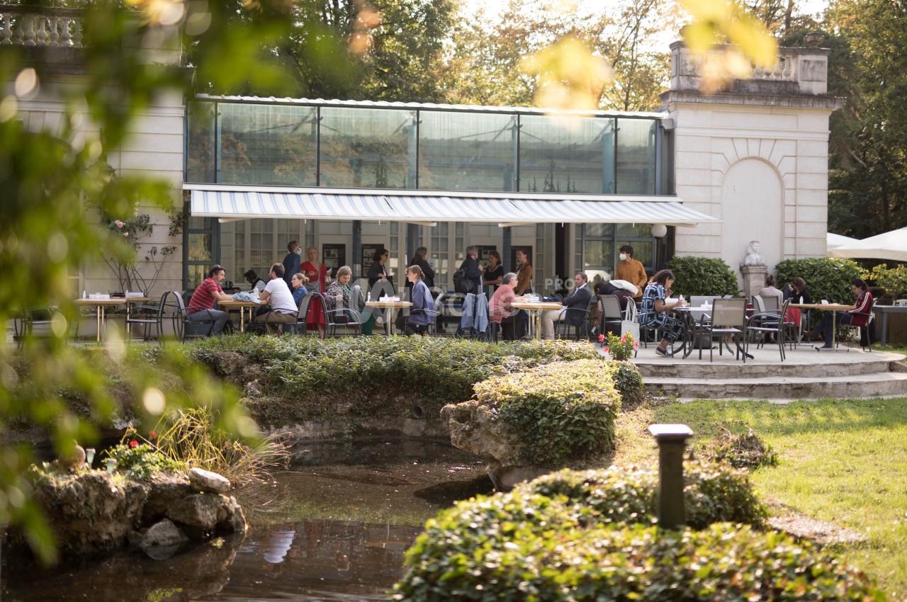 Terrasse d'un café animé près d'un petit étang, entourée de verdure et baignée par une lumière douce.