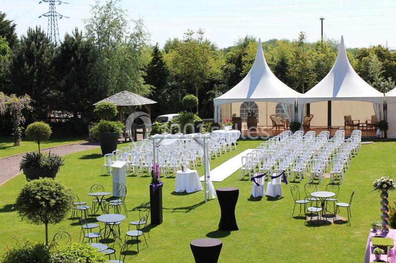 Chaises blanches alignées en extérieur pour une cérémonie, avec tentes blanches et tables noires dans un jardin verdoyant.