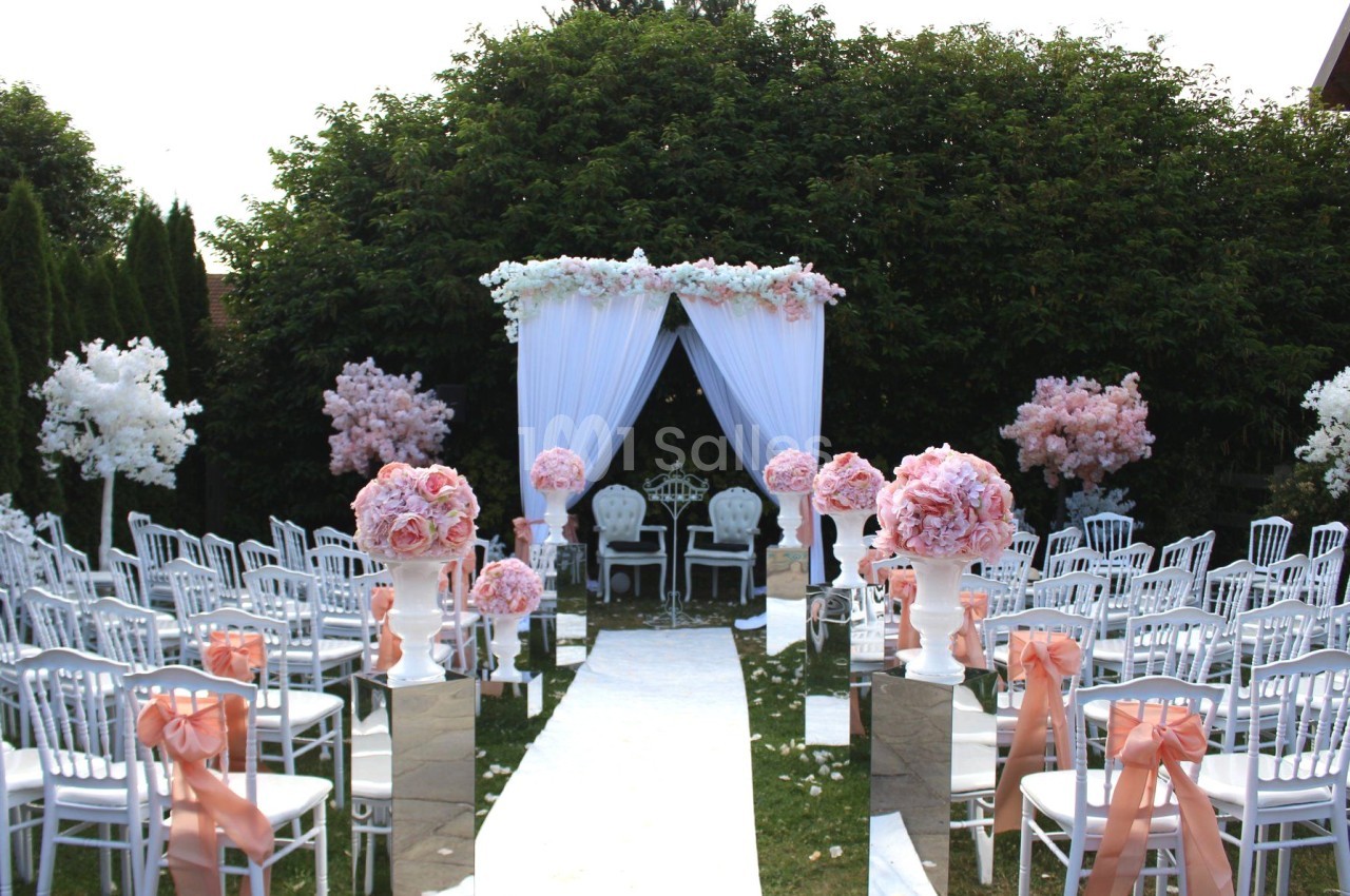 Allée centrale décorée de fleurs roses et blanches menant à une arche nuptiale dans un jardin extérieur.