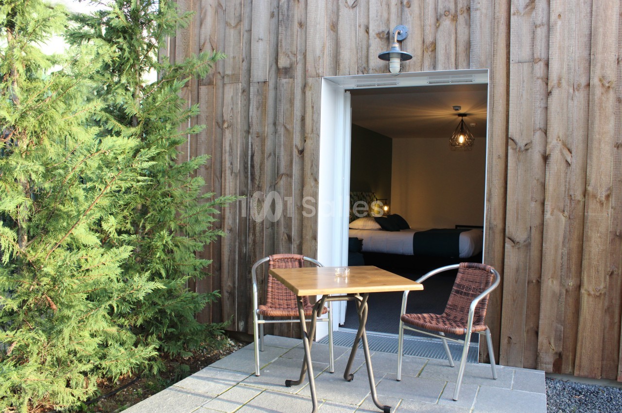 Terrasse avec table et chaises devant une porte ouverte donnant sur une chambre lumineuse, entourée de bois et de verdure.