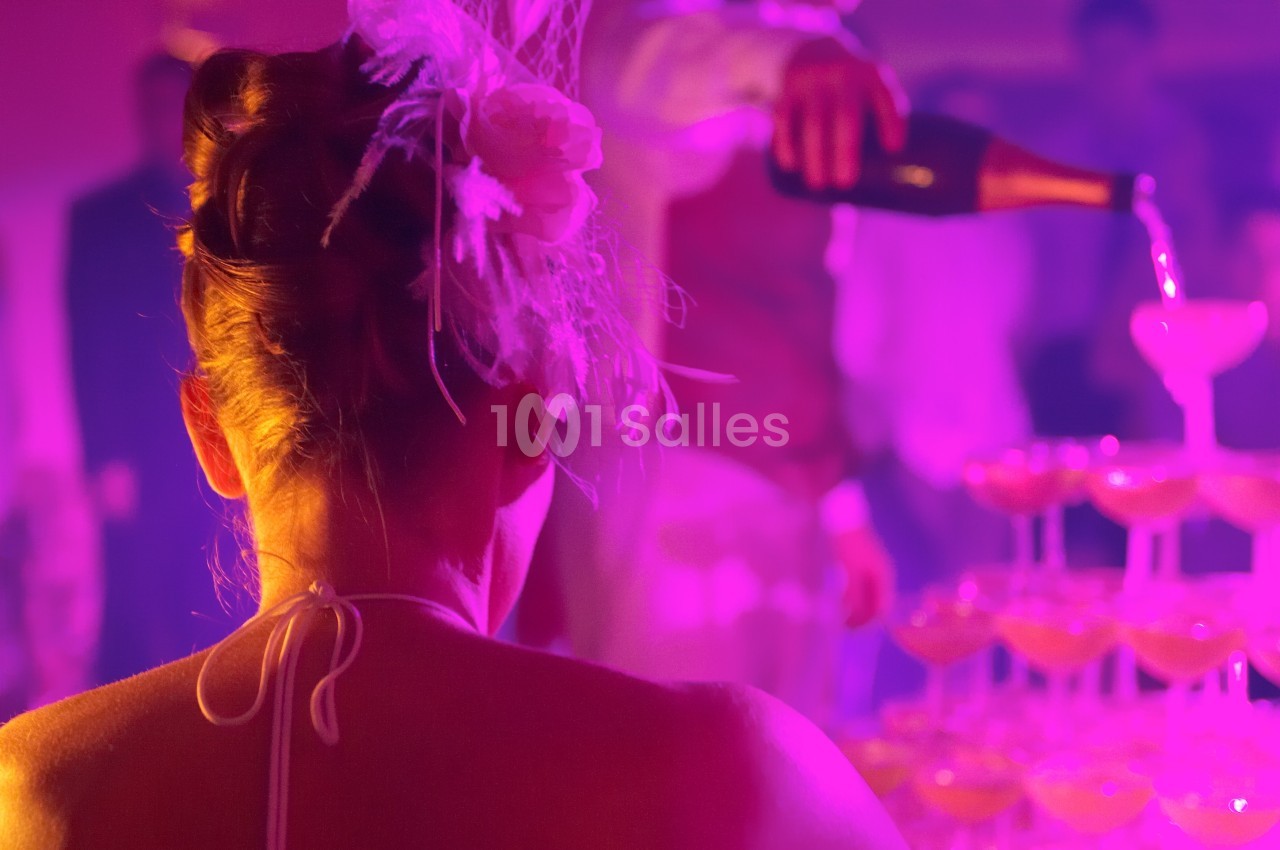 Une femme de dos observe un serveur verser du champagne dans une pyramide de coupes sous une lumière tamisée.