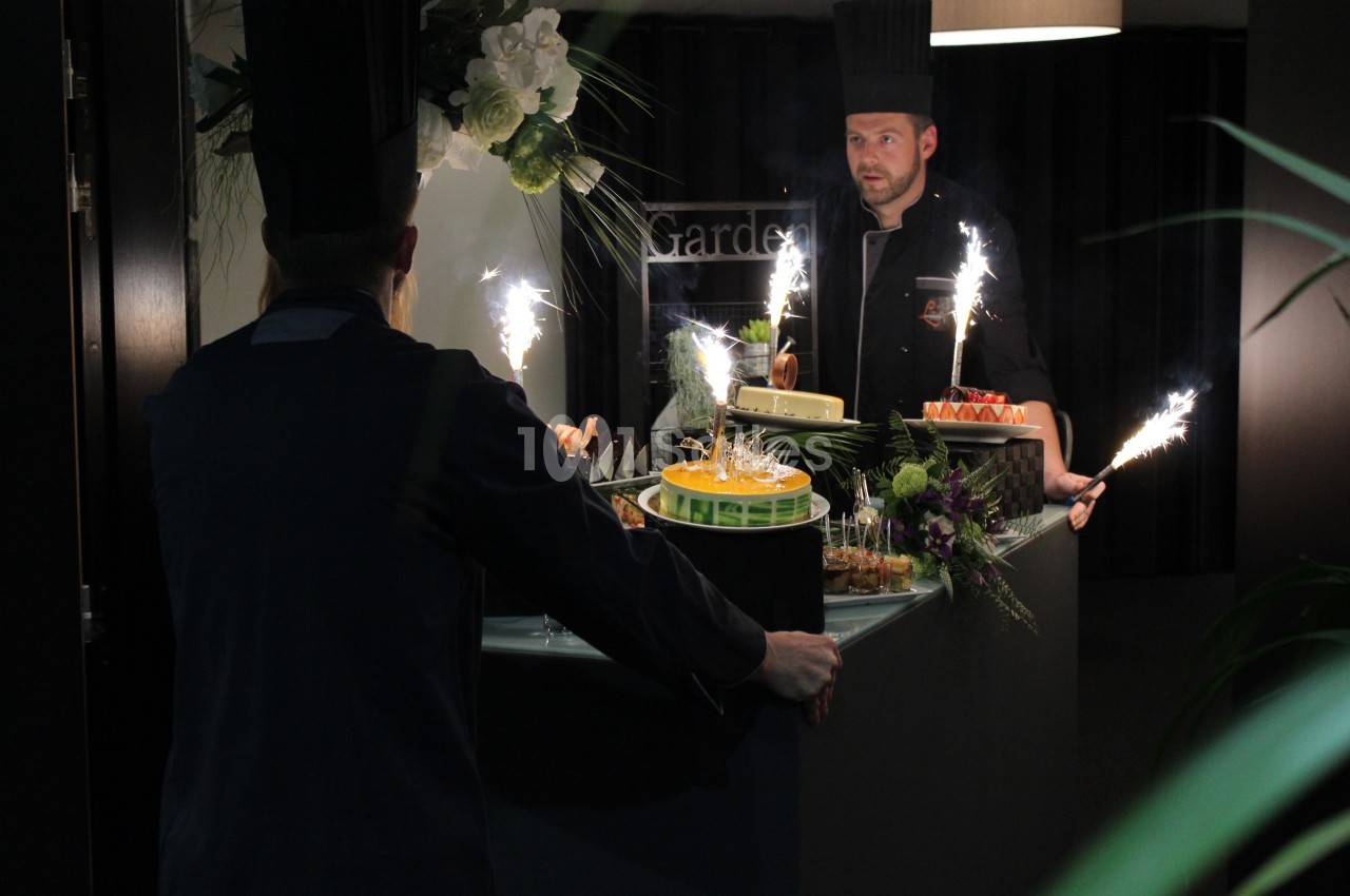Deux chefs en tenue présentent des desserts avec des cierges magiques sur un comptoir décoré de fleurs.