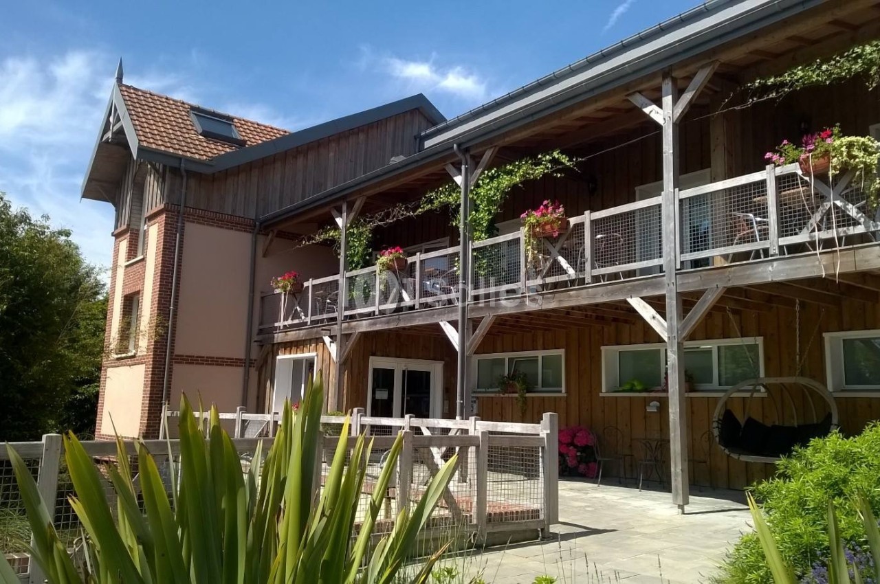 Bâtiment en bois avec balcon fleuri, grandes fenêtres et terrasse, entouré de verdure sous un ciel bleu.