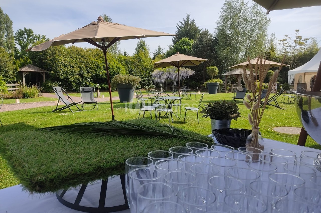 Table dressée avec des verres en premier plan, terrasse extérieure avec parasols, chaises et végétation en arrière-plan.