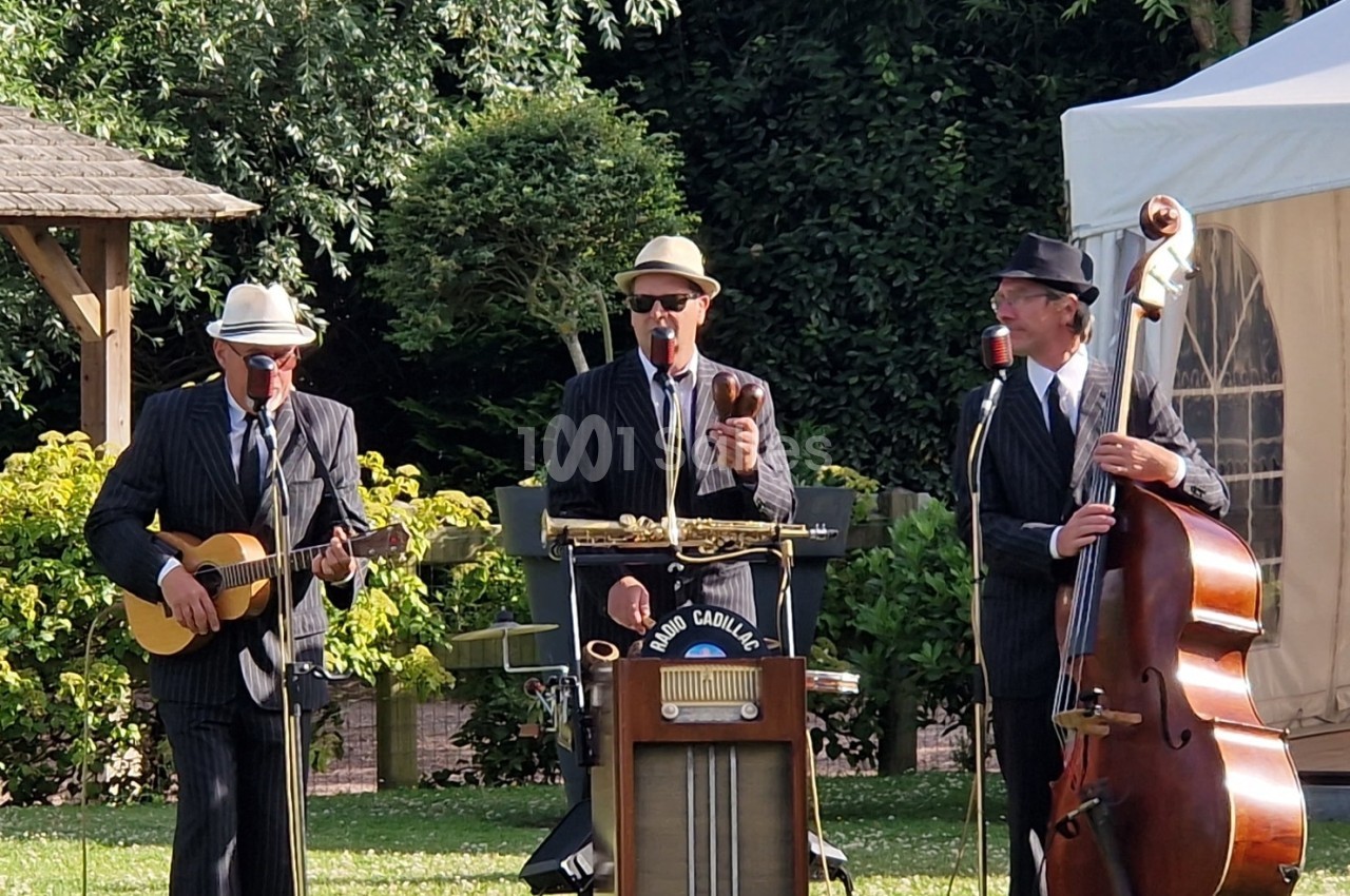 Trois musiciens en costume jouent de la guitare, du saxophone et de la contrebasse dans un jardin en plein air.