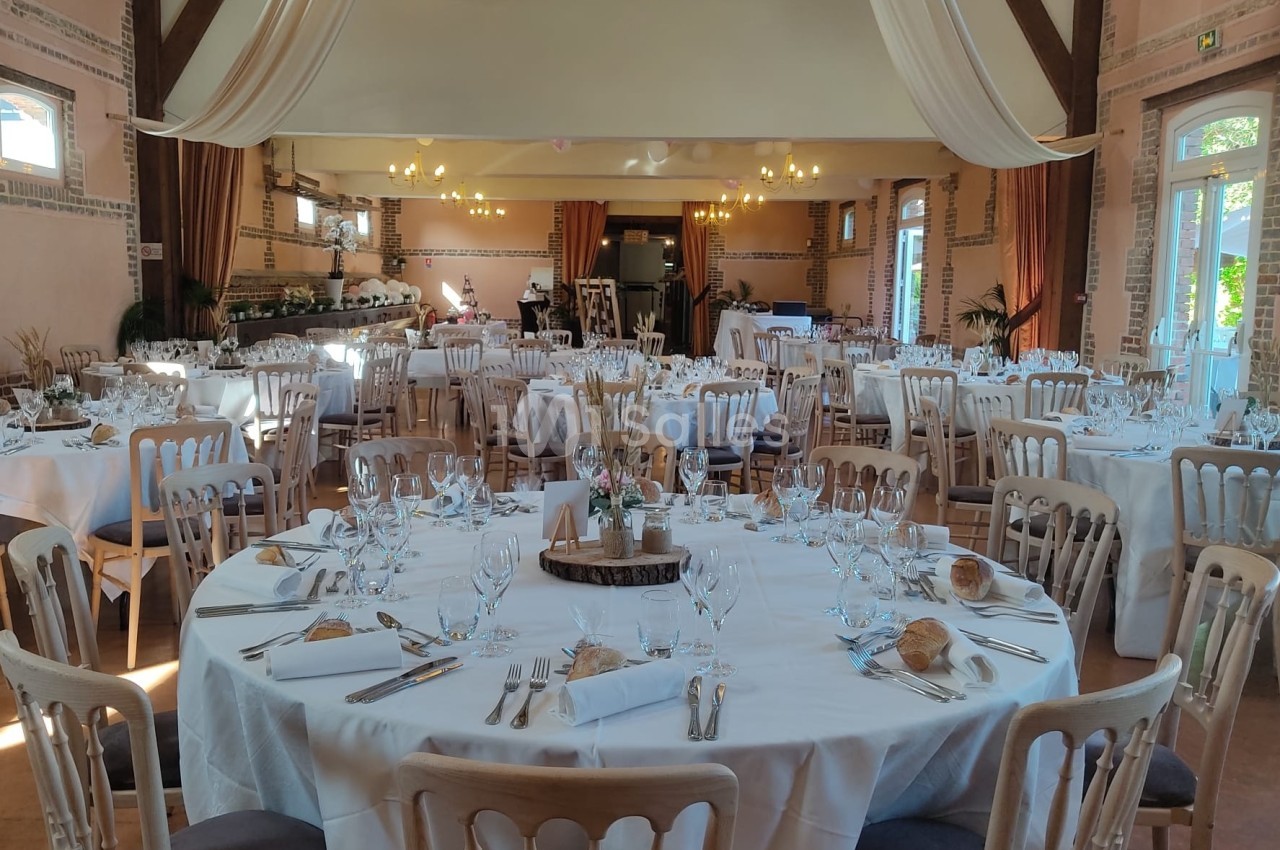 Salle de réception décorée avec des tables rondes dressées, nappes blanches et chaises en bois clair.
