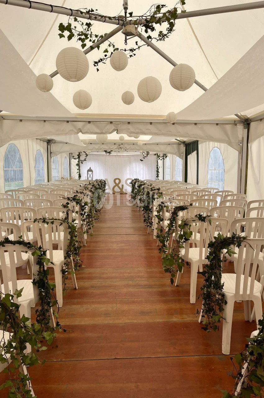 Allée centrale d'une tente de mariage décorée de guirlandes de lierre, avec des chaises blanches et des lanternes suspendues.