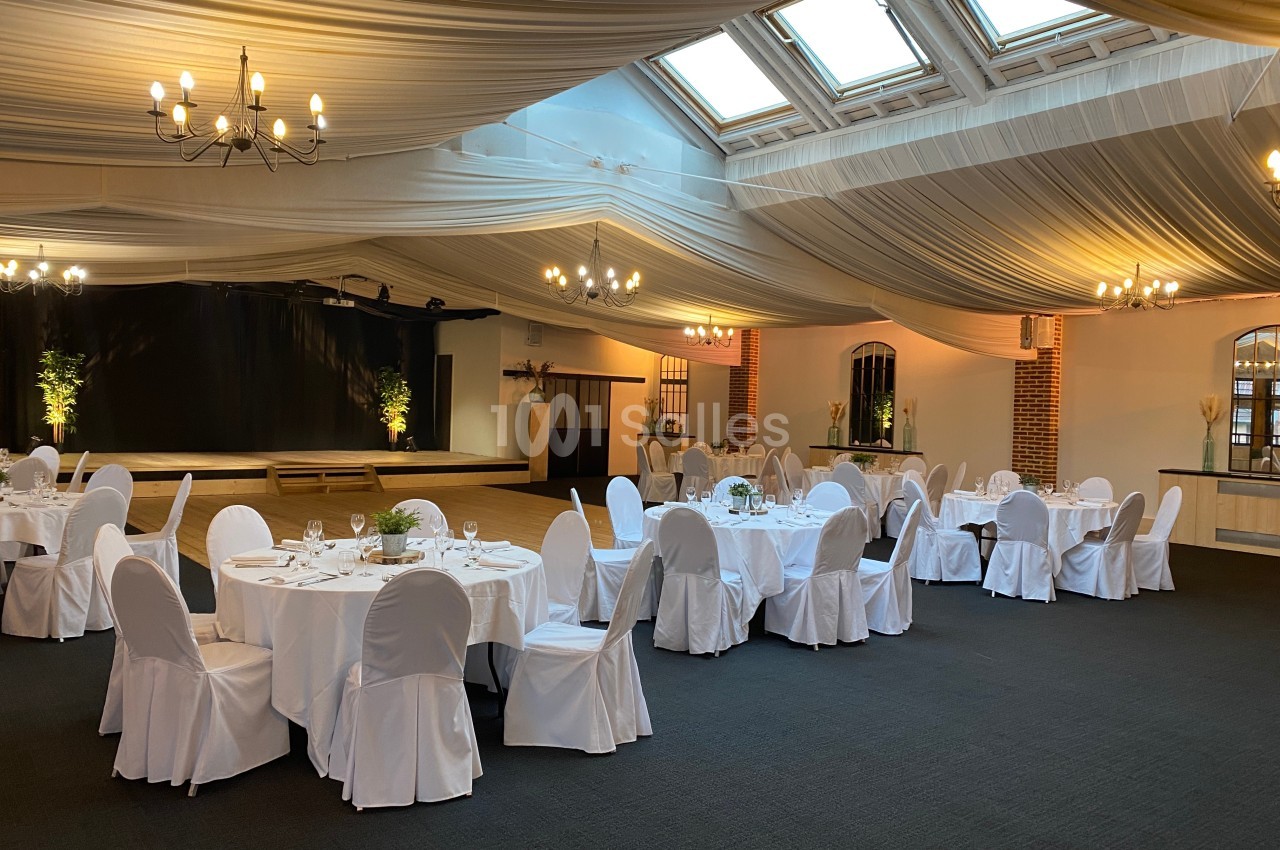 Salle de réception élégante avec tables rondes dressées, chaises blanches et plafond drapé sous une lumière naturelle.
