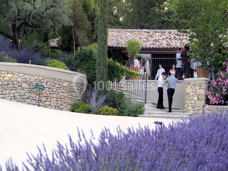 Maison en pierre entourée de lavande et de végétation, avec des personnes discutant près d'une terrasse.